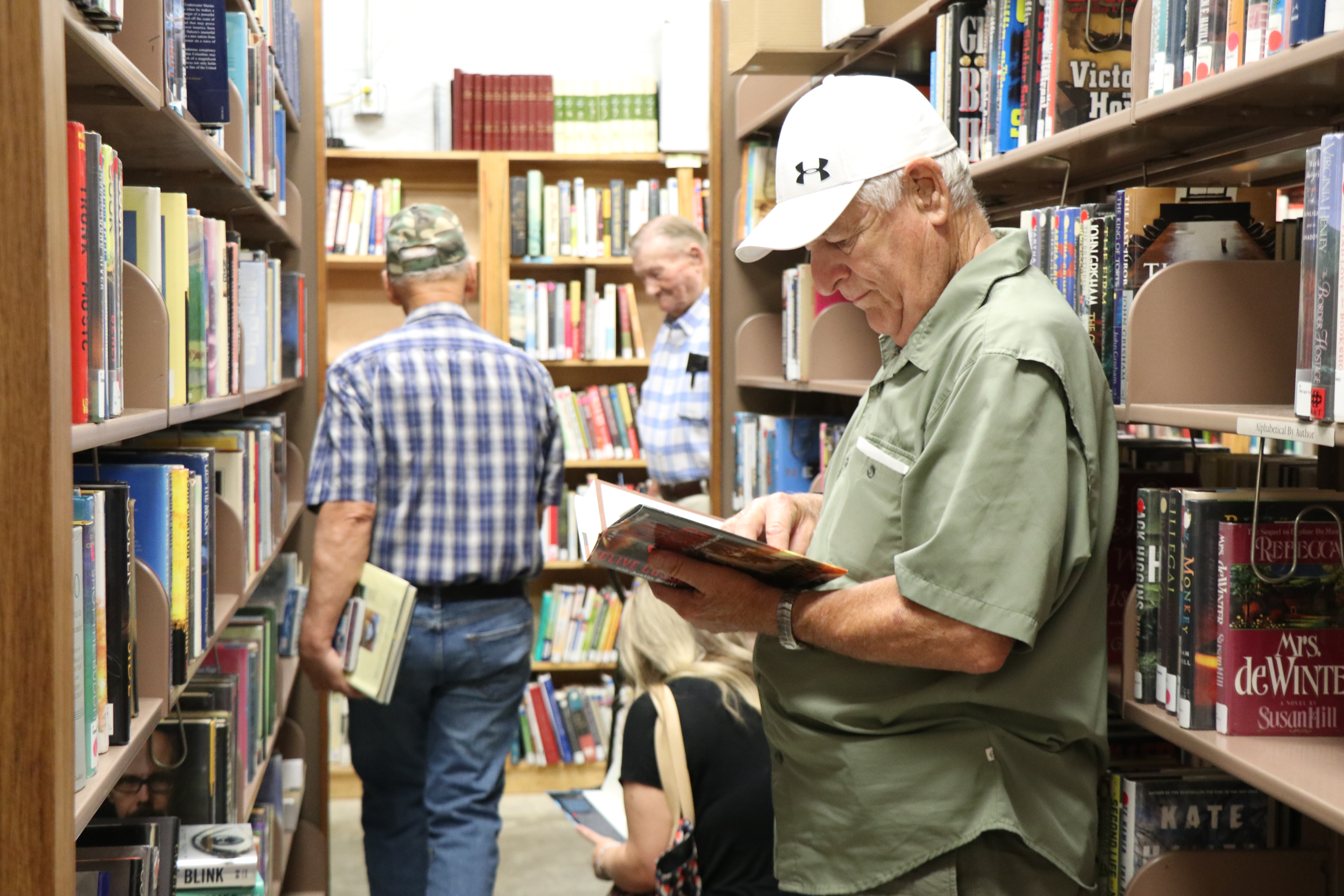 Two men stand in between bookshelves. One is looking down at a book. 