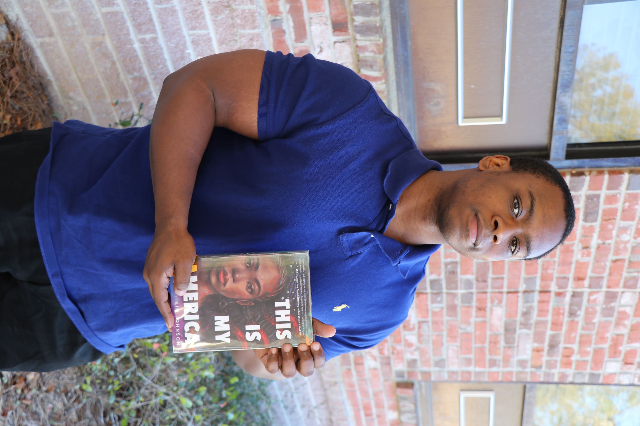 A man stands in front of a brick wall. He is holding the book This Is My America.