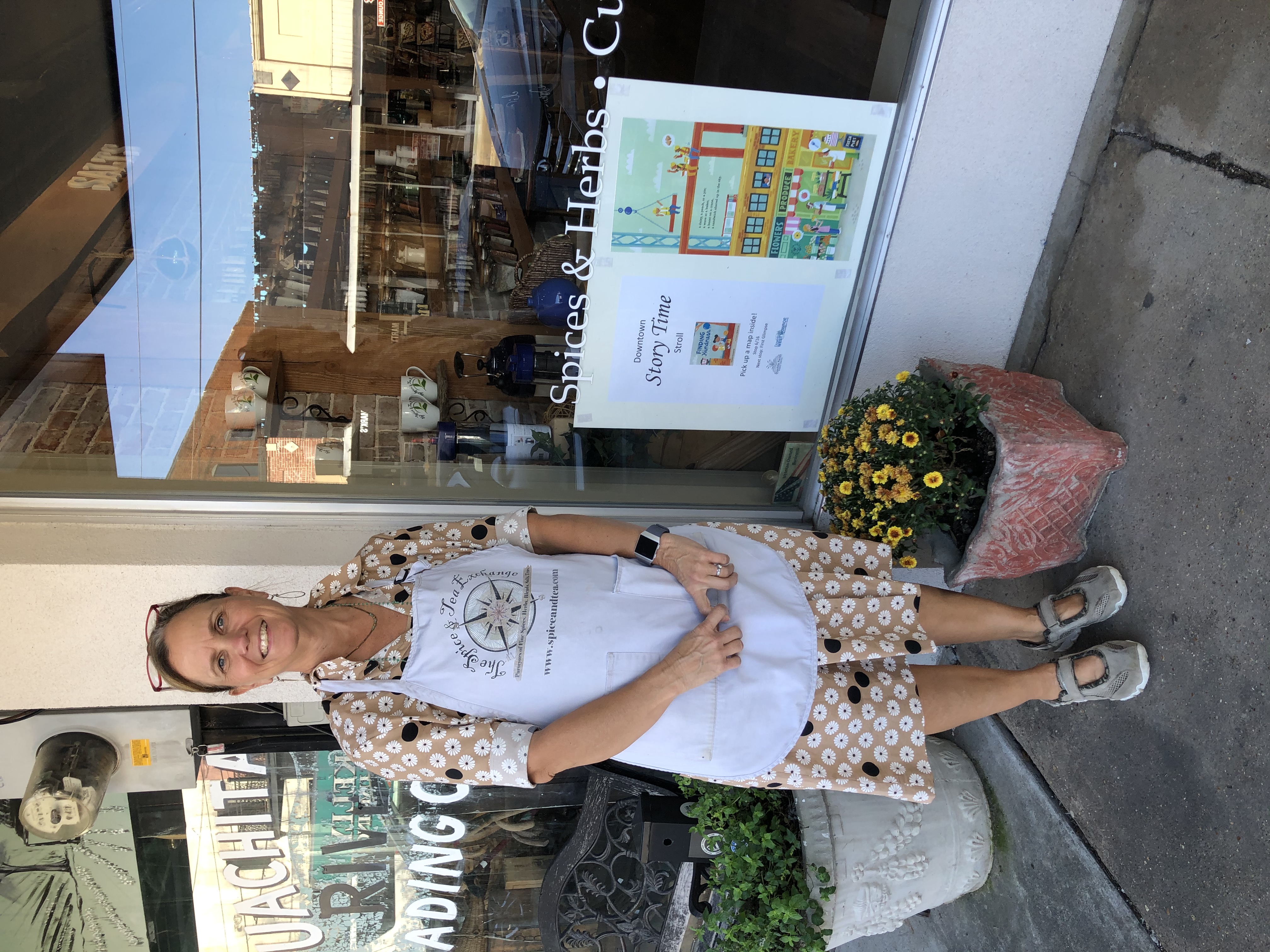 Woman in apron stands outside a shop window. A page from the book Finding Kindess hangs in the window.