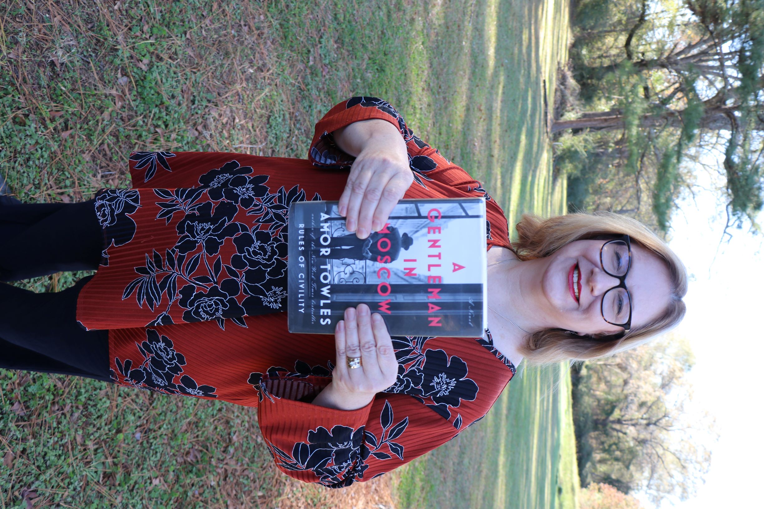 A woman stands in a field. She is holding the book A Gentleman in Moscow.