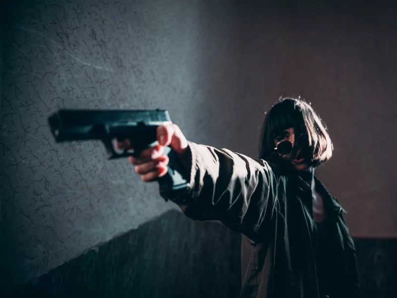 In a stairwell lit only by a window behind her, a woman looks over her sunglasses to aim a pistol