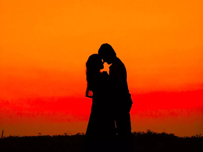 A woman and man stand together, foreheads and noses touching, silhouetted against an orange sunset streaked with red