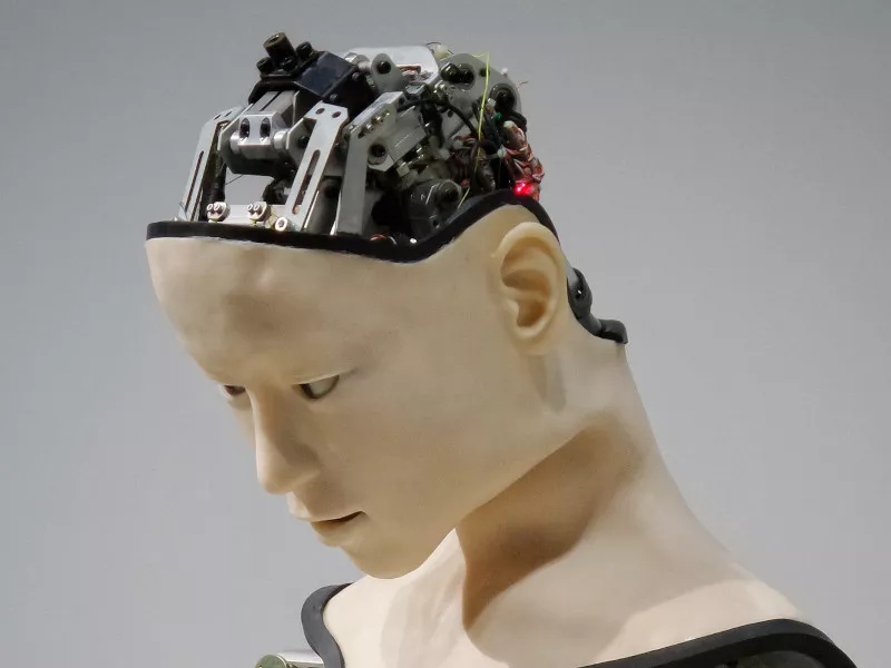 Robotic parts including wires and a camera are visible inside the opened skull of a robot with a human shape