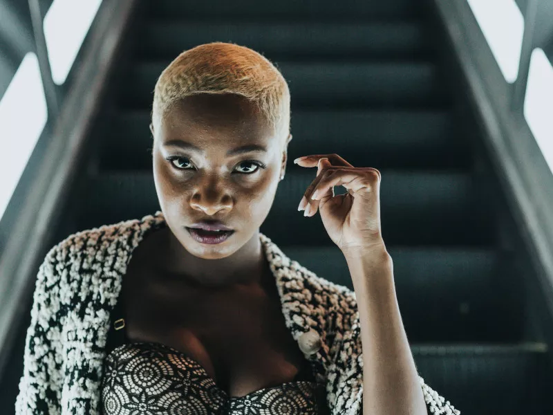 A Black woman with short bleached hair regards the viewer as she sits on an outdoor escalator which lights her face