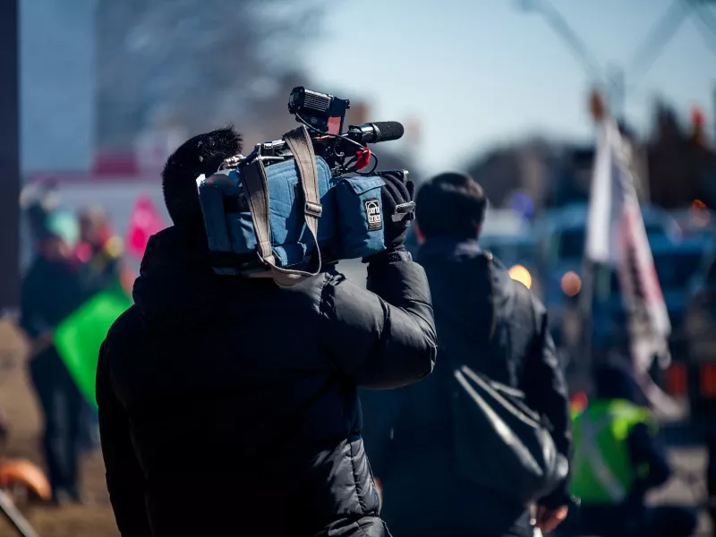 A camera operator stands in front of a scene with first responders wearing safety yellow vests