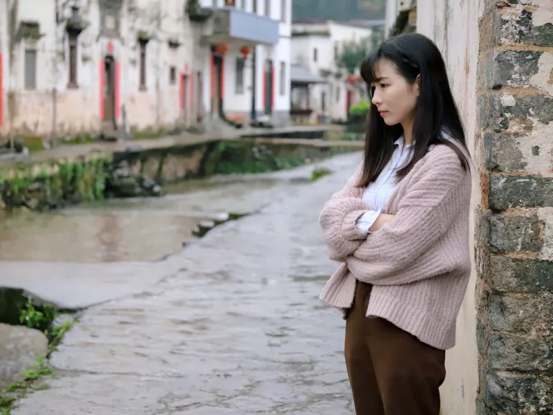 A woman, crossing her arms unhappily, leans against a building in front of an urban canal