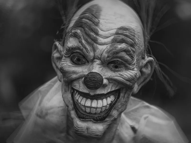 A gray clown mask with alligator eyes and huge eyebrows leers with a toothy grin.