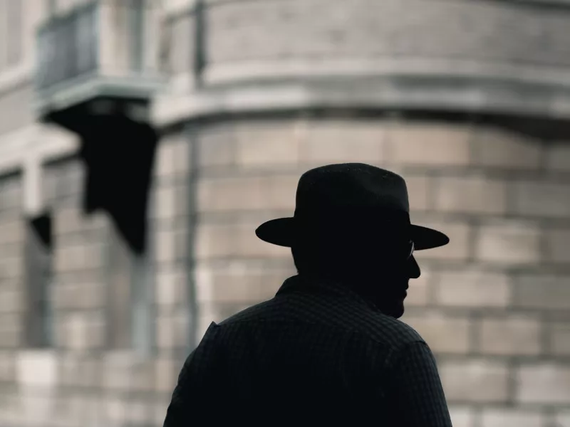 A silhouette of a man with a hat standing in the shadows near the corner of a building