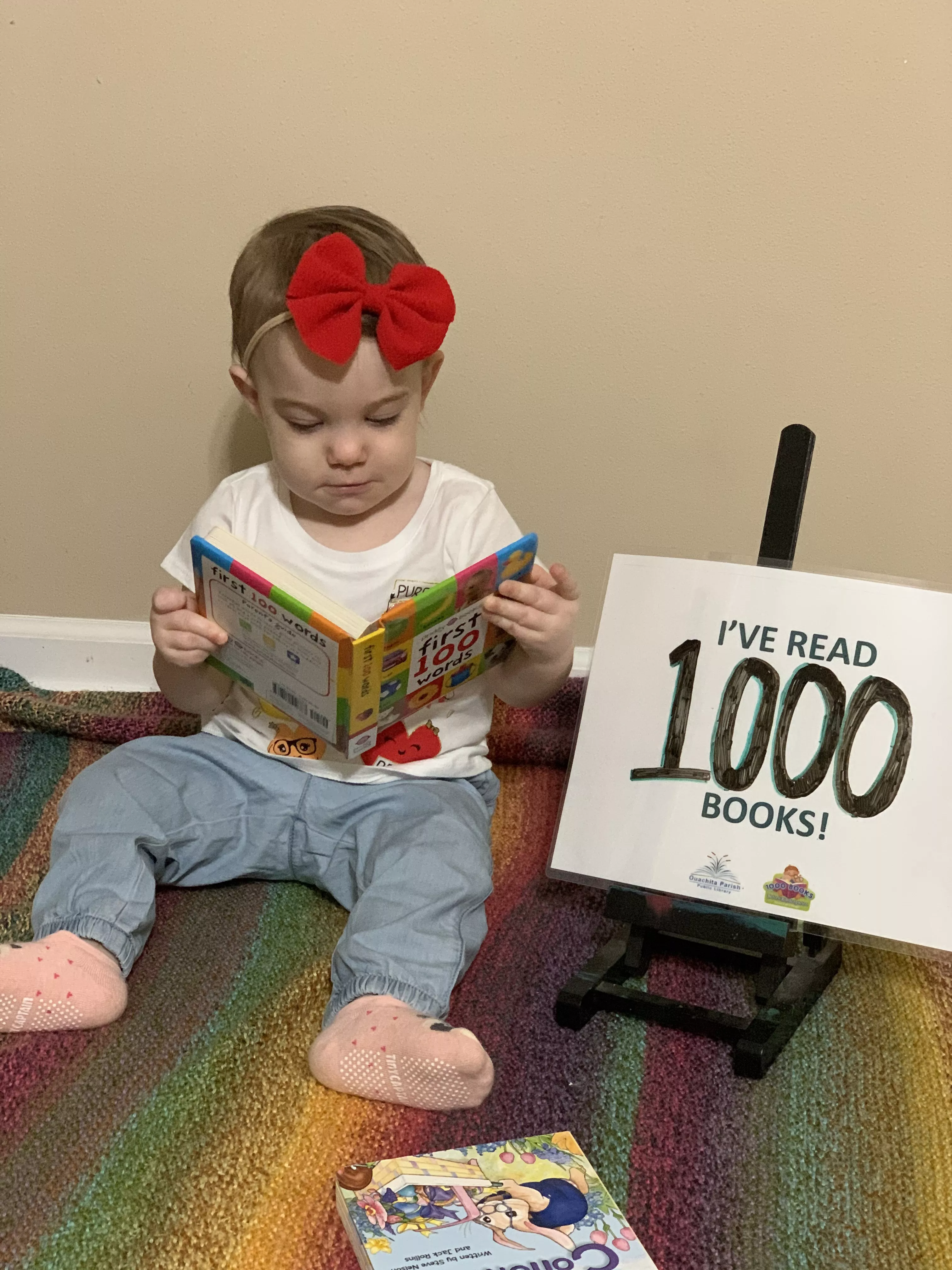 A baby sits up with a book open in her lap. There is a sign next to her that says, &quot;I&#039;ve read 1000 books!&quot;