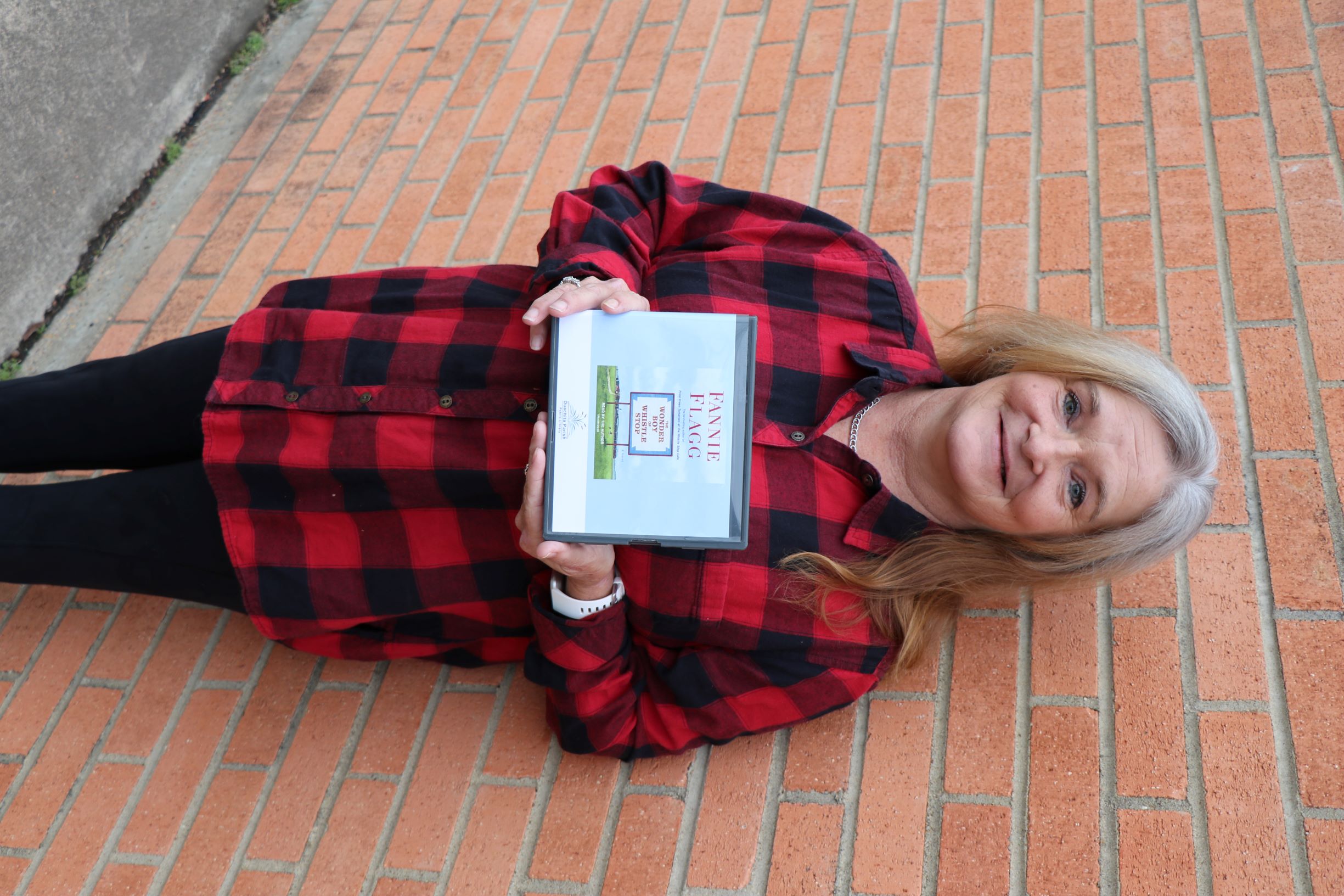 a woman stands in front of a brick wall. She is holding the book-on-CD, The Wonder Boy of Whistle Stop by Fannie Flagg.